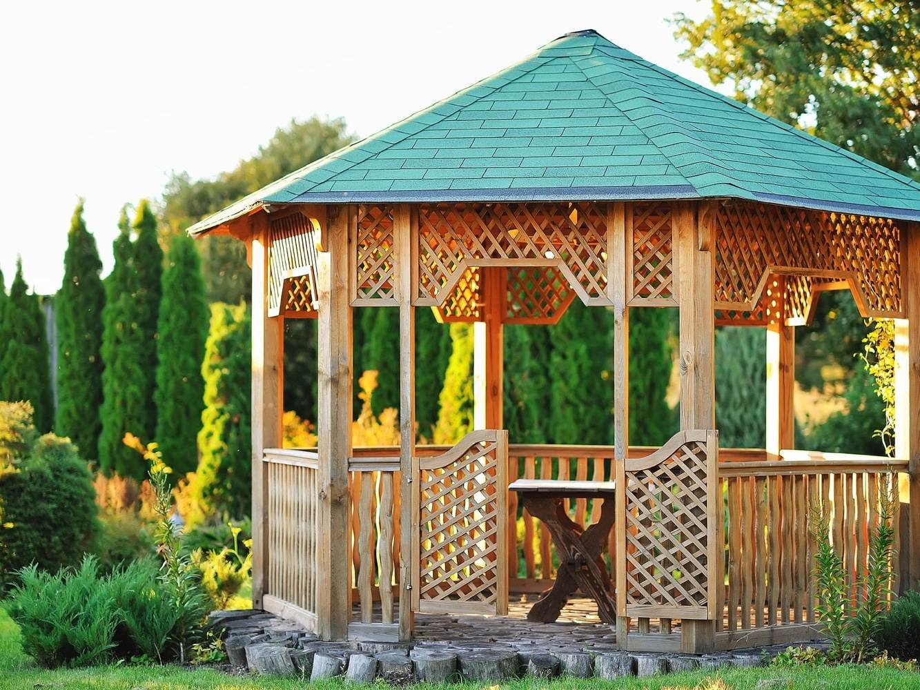 This image shows a beautiful gazebo with a green roof with pine trees behind it and the sun shining.