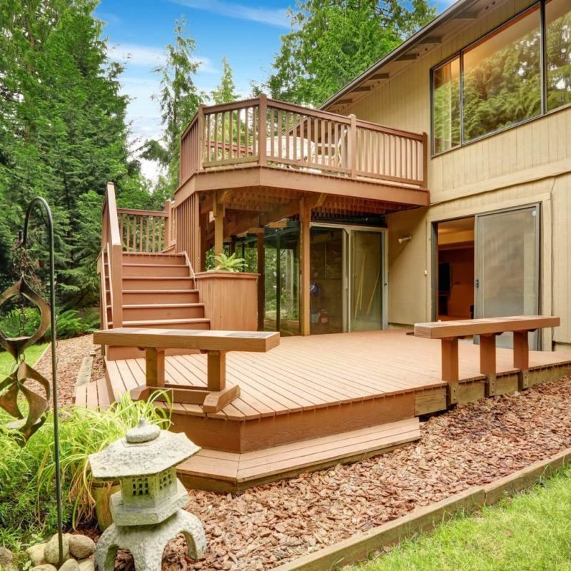This image shows a new two level deck with stairs attached to a brown house with pine trees in the background and professional landscaping done in North Carolina.